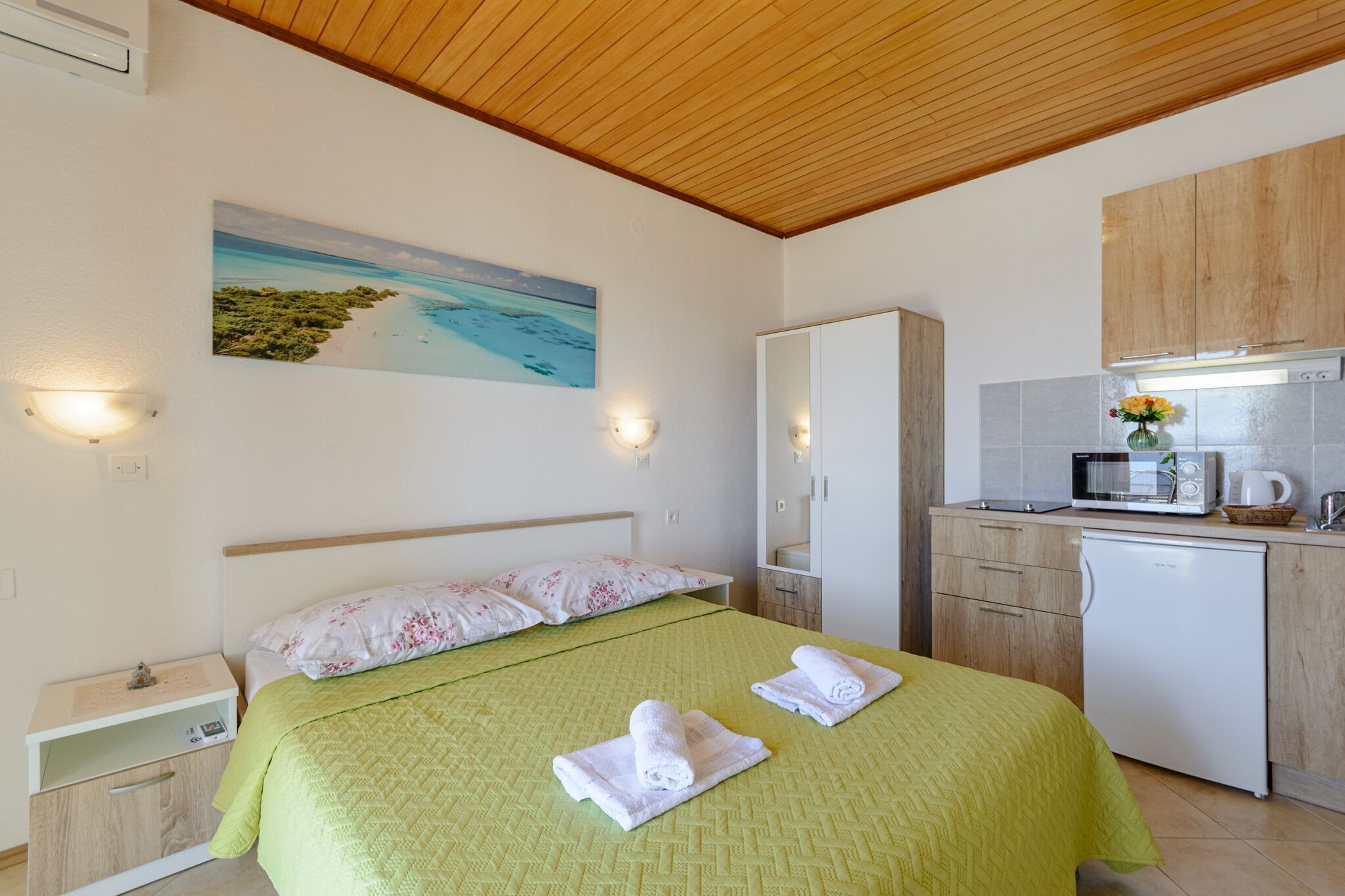 A bedroom with a bed, a nightstand, a bedside table, a wall-mounted light, and a painting of a beach scene.