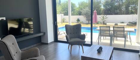 A modern living room with a TV, a sofa, a dining table, and a view of a swimming pool.