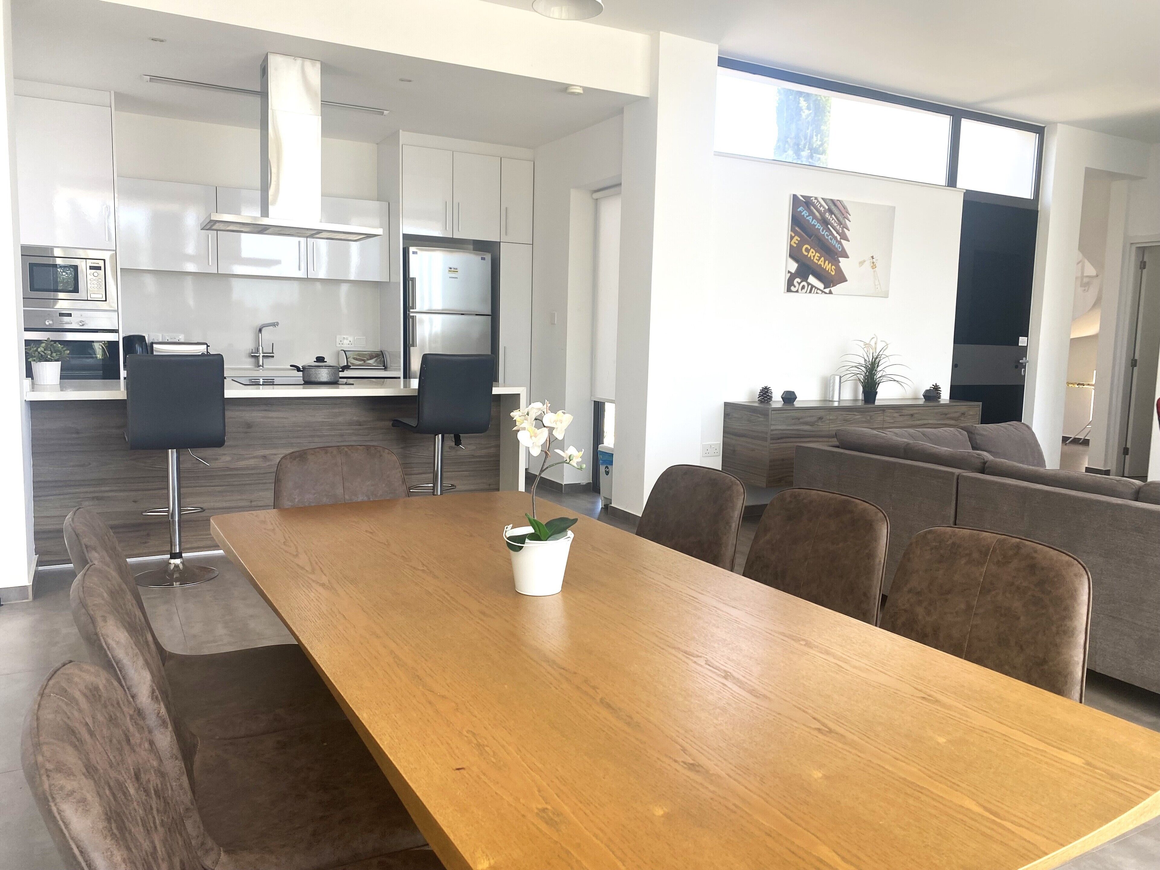 A modern kitchen with white cabinets, a stainless steel refrigerator, and a microwave. There is a dining area with a wooden table and brown chairs.