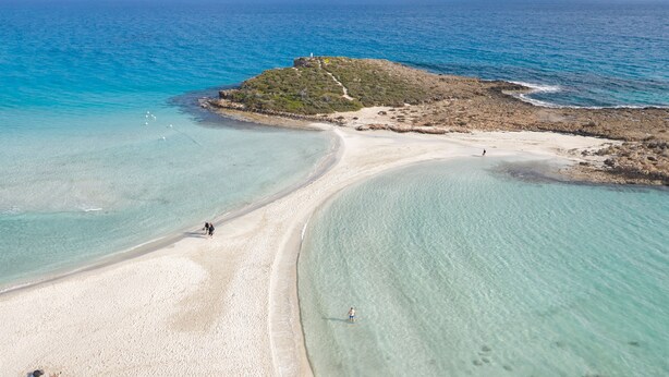 Μια αεροφωτογραφία μιας παραλίας με κρυστάλλινα, γαλαζοπράσινα νερά, μια αμμώδη ακτή και έναν μικρό λόφο με ένα κτίσμα στην κορυφή.