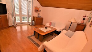 Un salón acogedor con piso de madera, un sofá, una mesa de centro con un vaso decorativo, un televisor sobre un mueble de madera y un ventanal corredizo que da a un balcón.
