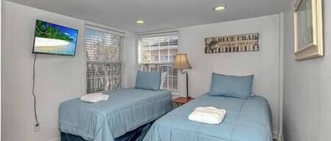 A bedroom with two single beds, a TV on the wall, and a window with blinds.
