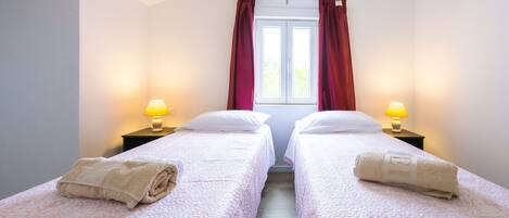 Een hotelkamer met twee eenpersoonsbedden, elk voorzien van een kussen en een opgerolde handdoek. Aan weerszijden van de bedden staan nachtlampjes.