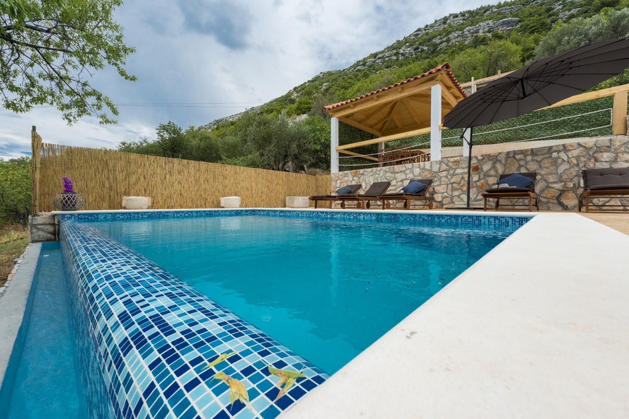 A swimming pool with tiled edges, lounge chairs, a covered seating area, and a stone wall.