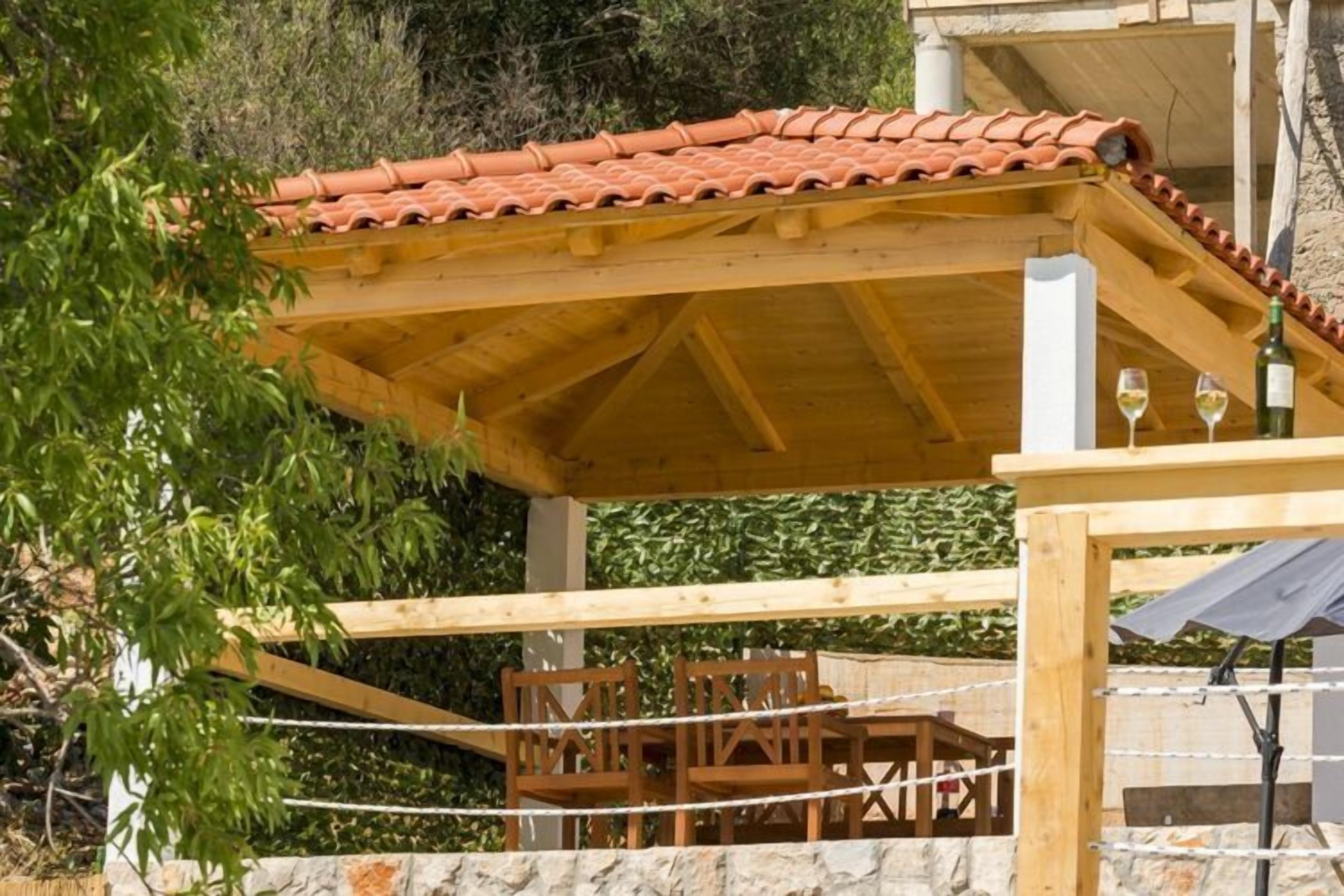 En træpergola med tegltag, stenvægge og et bord med stole under.