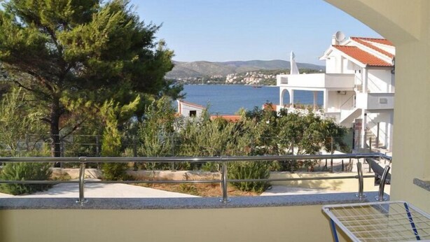 Un balcón con vista al mar, un árbol y edificios al fondo.