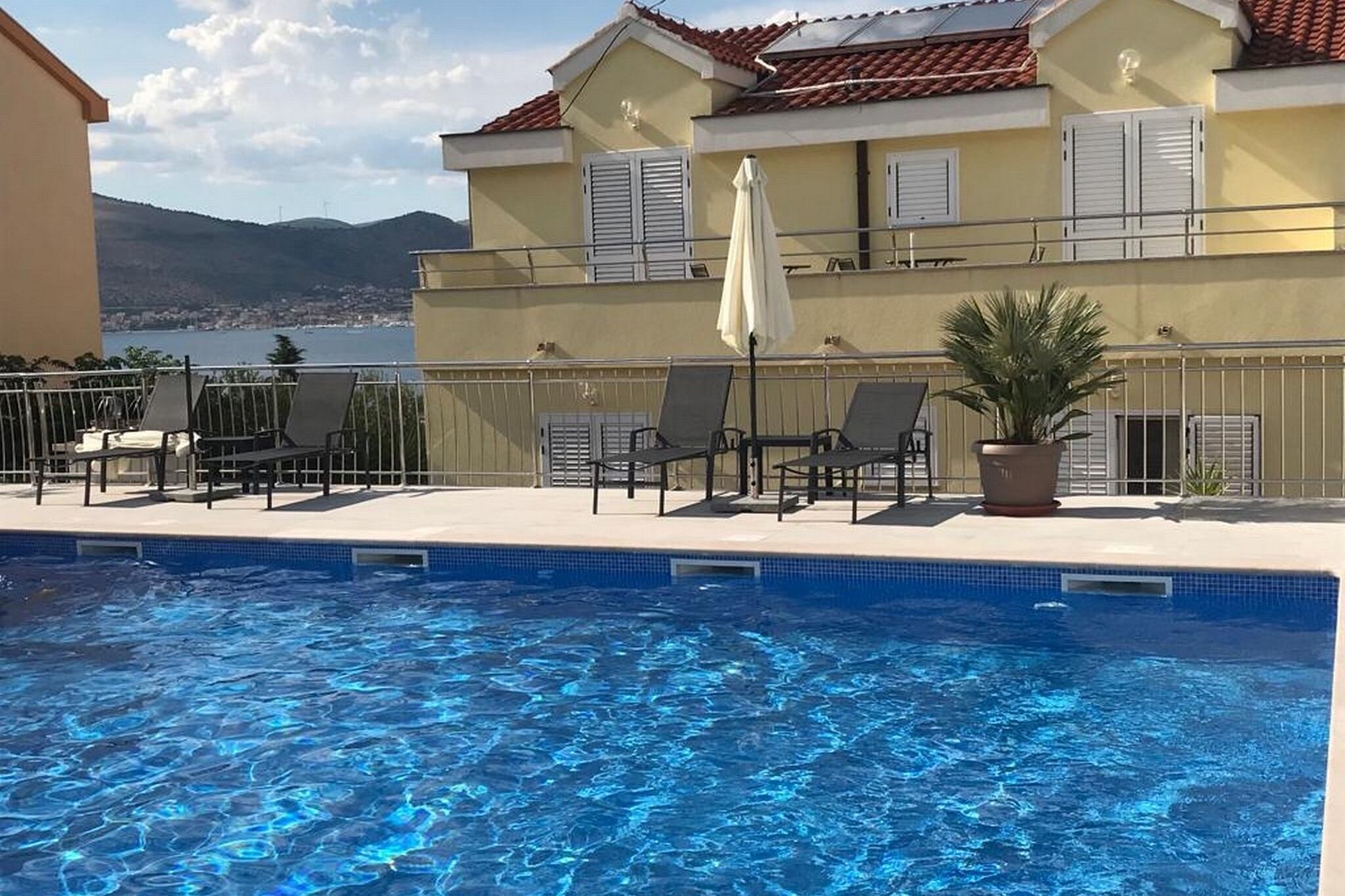 Una piscina con sillas de descanso y una sombrilla, rodeada por un edificio con techo de tejas y vistas al mar.