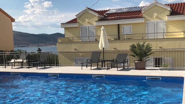 Una piscina con sillas de descanso y una planta en maceta, un balcón con barandilla, y un edificio con tejado de tejas rojas y contraventanas blancas.