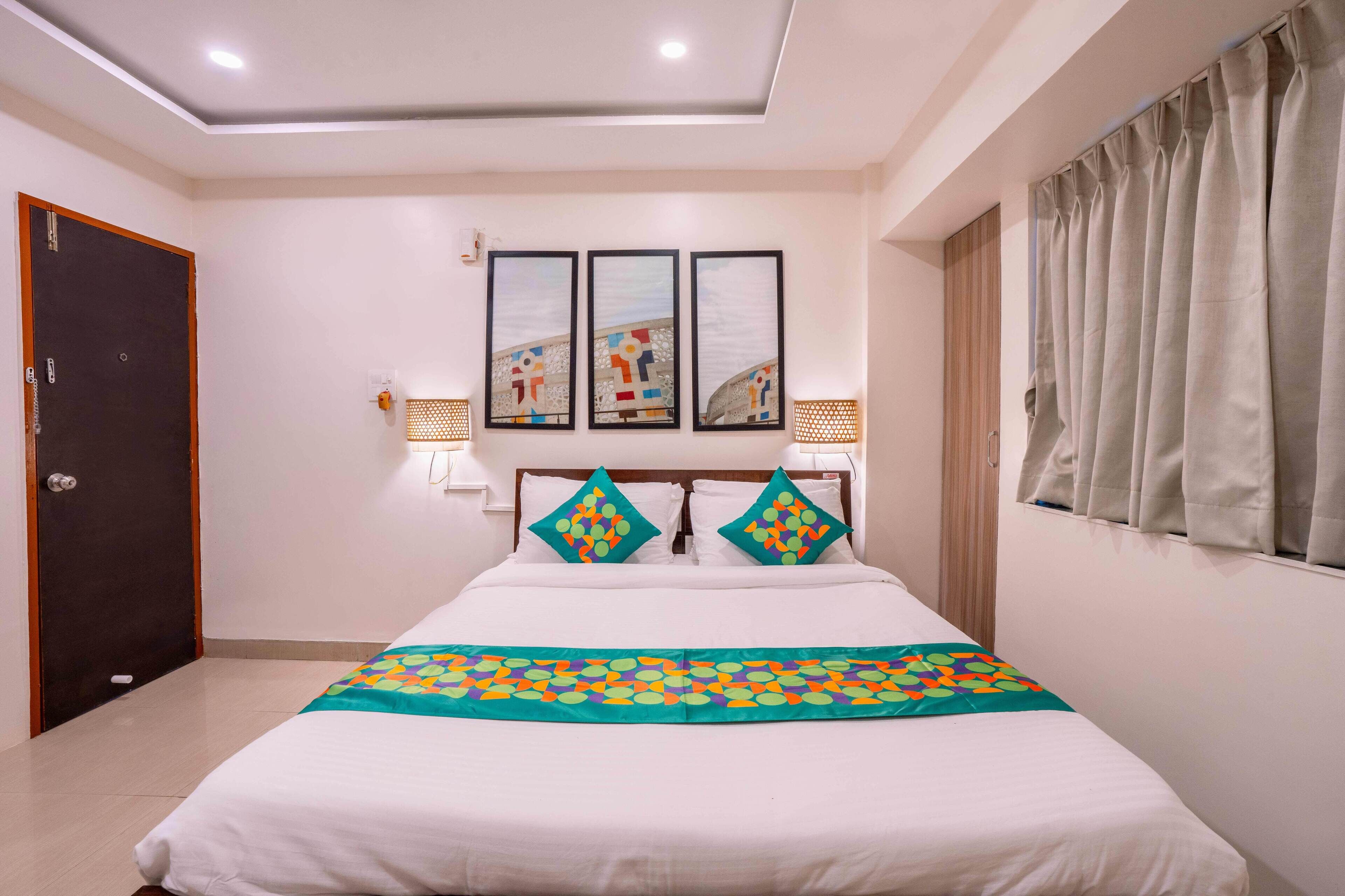 A hotel room with a bed, a door, a window with curtains, and wall-mounted artwork.