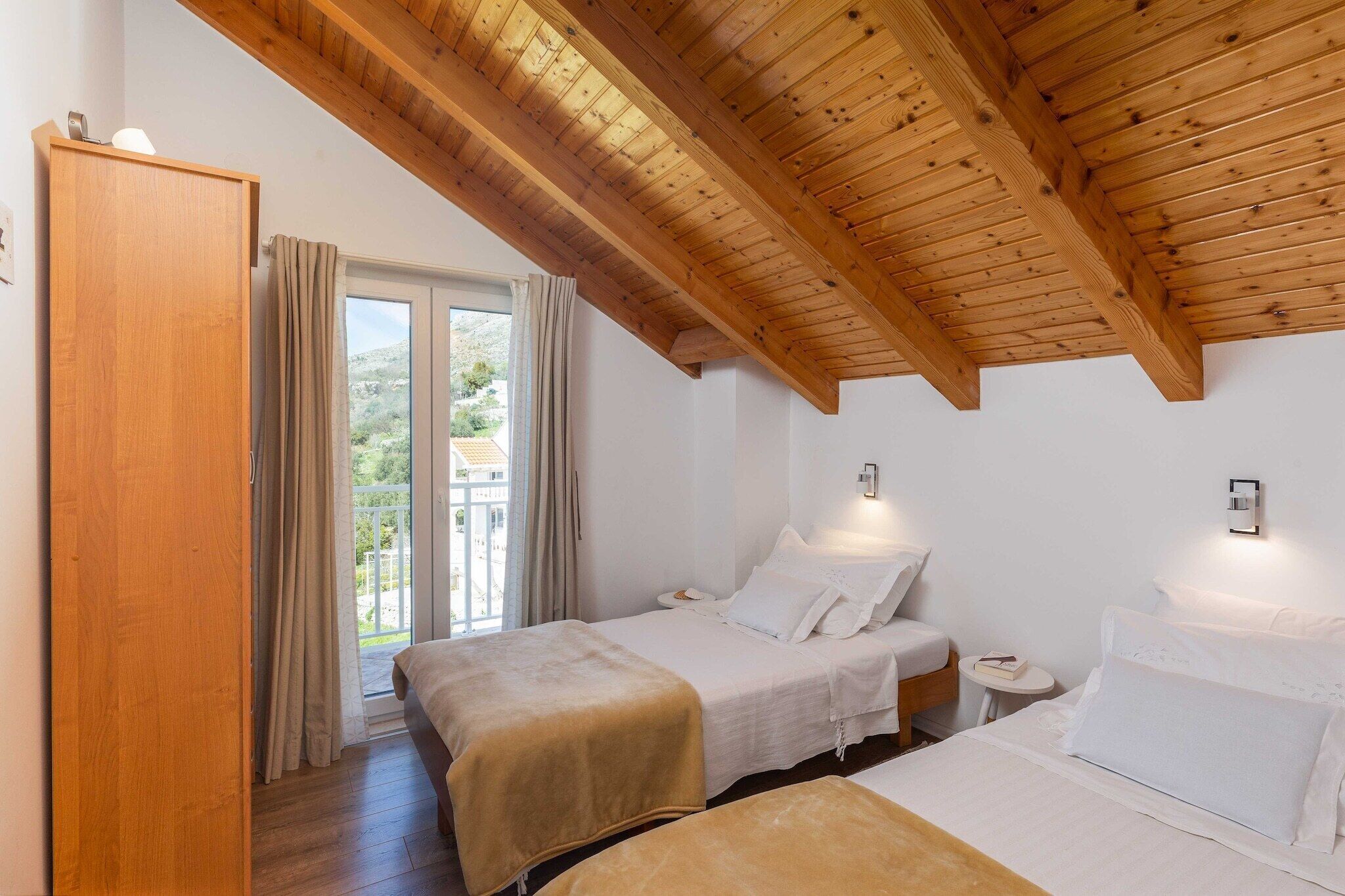 Une chambre avec deux lits, des poutres apparentes en bois au plafond, une armoire et une fenêtre avec des rideaux.