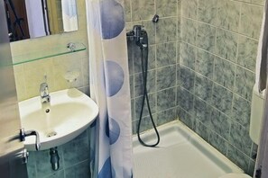 A bathroom with a white sink, a glass shelf, a shower with a handheld hose, and tiled walls.