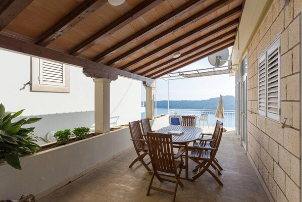 A covered outdoor patio with a table and chairs, a view of the sea, and a building with shutters. - Apartments Sipa - Superior Two Bedroom Apartment with Balcony and Sea View (Apartman F) (Dubrovnik)