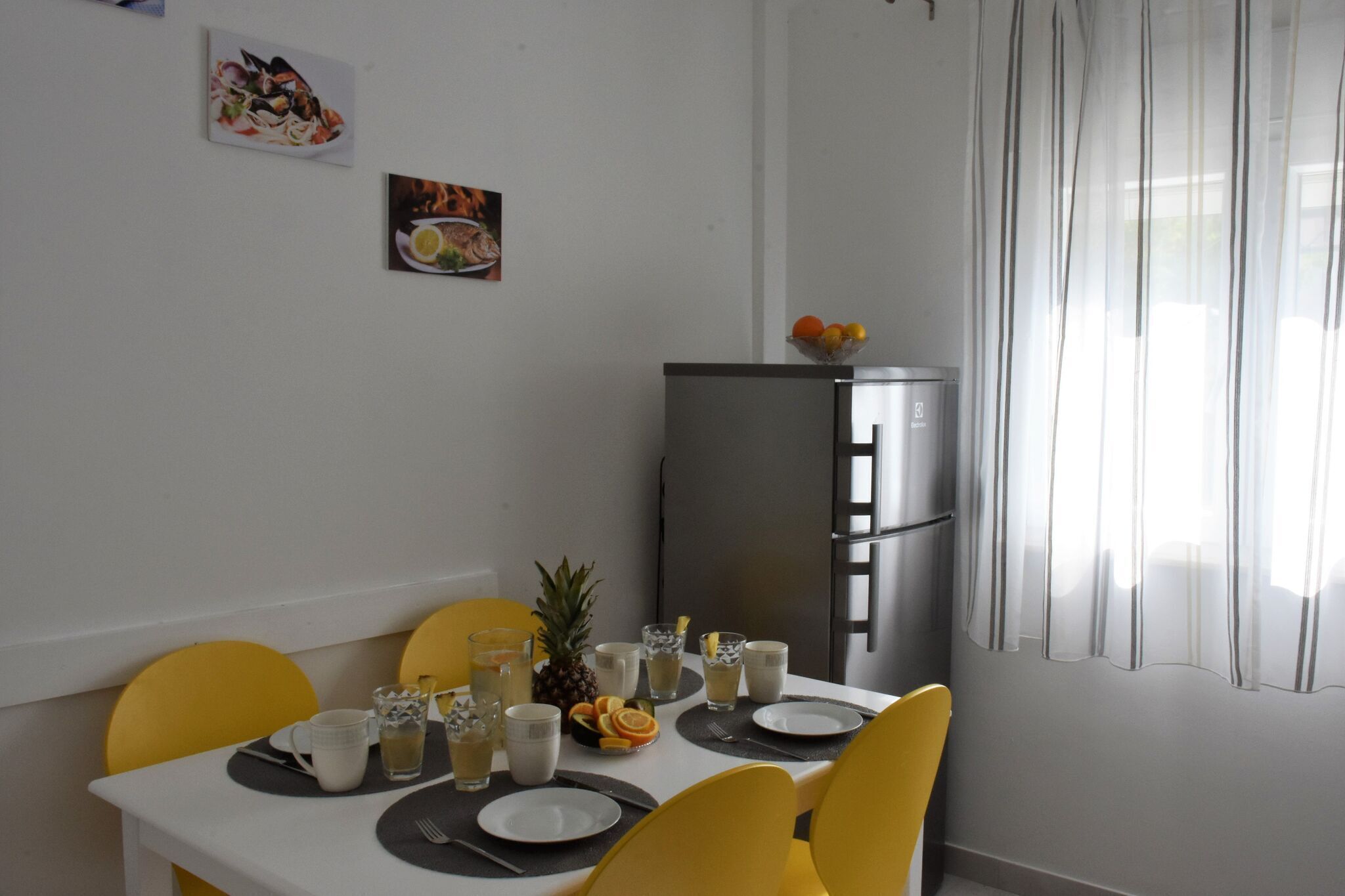 Ein Essbereich mit einem weißen Tisch, gedeckt für vier Personen, gelben Stühlen, einem Kühlschrank und zwei Wandbildern mit Speisenmotiven.