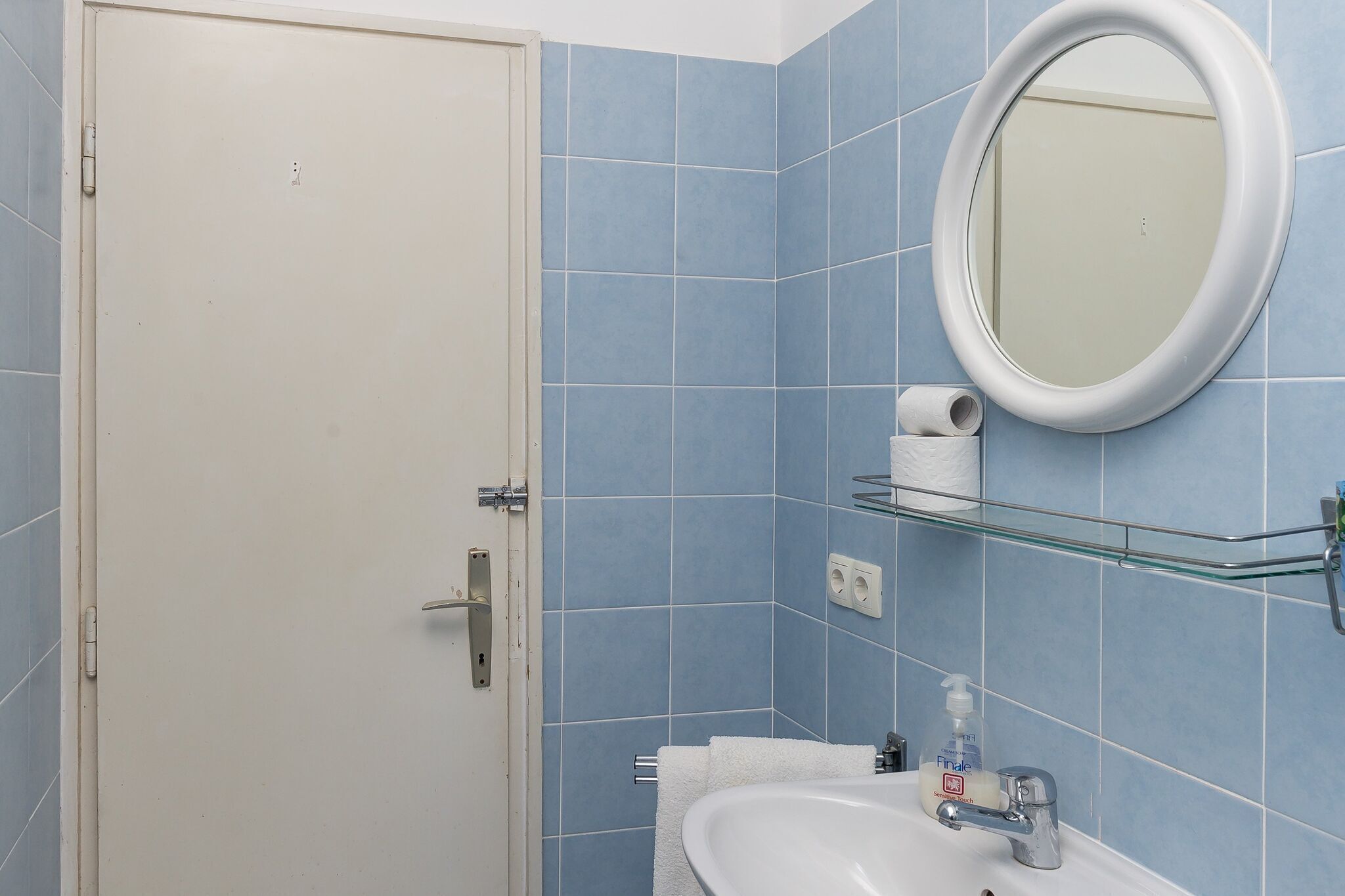 A bathroom with blue tiles, a white door, a round mirror, a glass shelf, a sink, and a toilet paper holder.