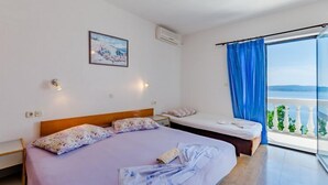 A bedroom with a bed, a bedside table, a nightstand, a window with blue curtains, and a view of the sea. - Villa Kosa - Two Bedroom Apartment with Balcony and Sea View (A2 Plavi) (Brela)