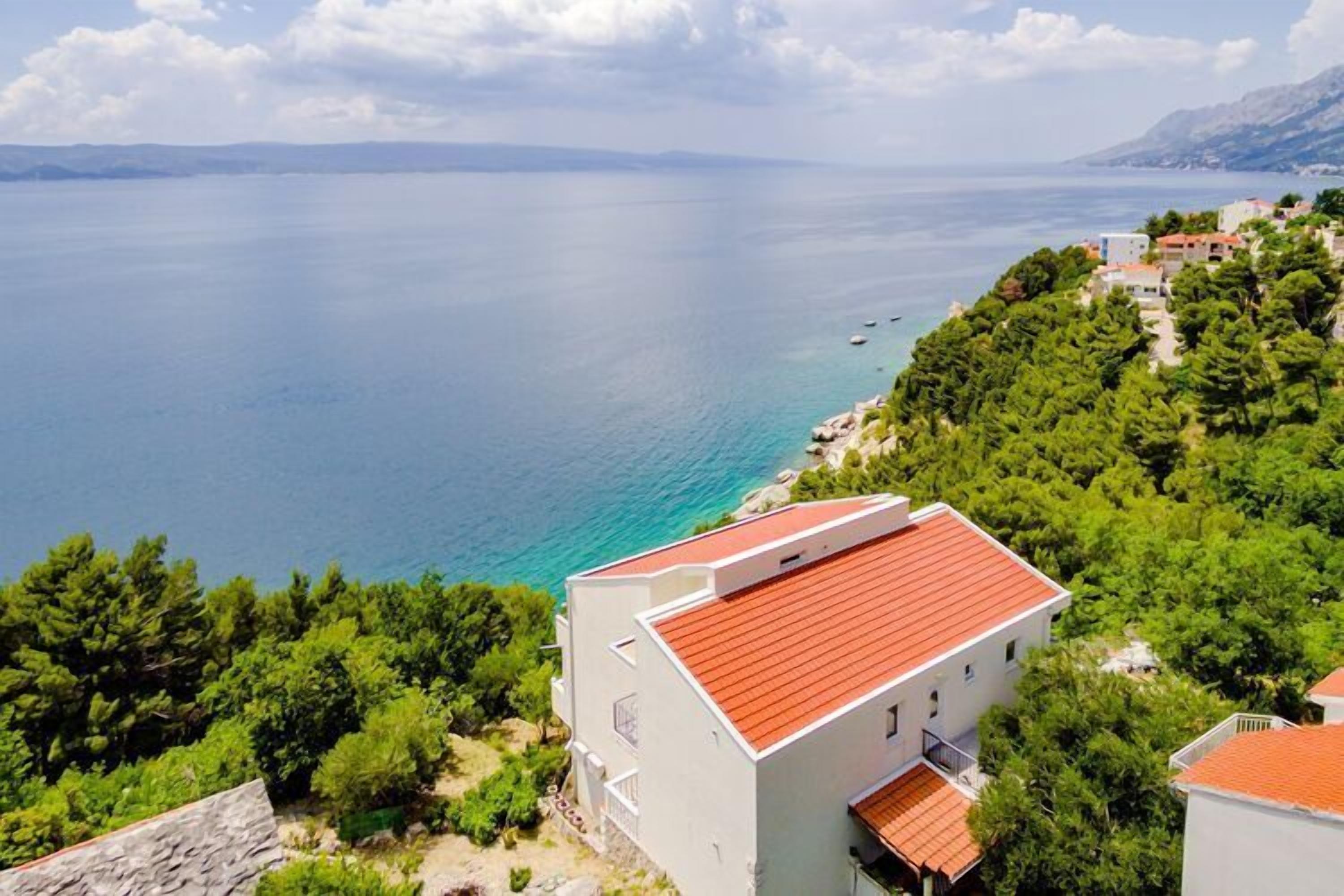 Un edificio costero con techo rojo, rodeado de árboles verdes frondosos y vista al mar.