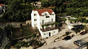 A white two-story building with a red-tiled roof, surrounded by greenery, with a parking area and a small garden in front. - Apartments Cypress Sea Cove - Comfort Studio Apartment with Balcony and Sea View (A1) (Slano)