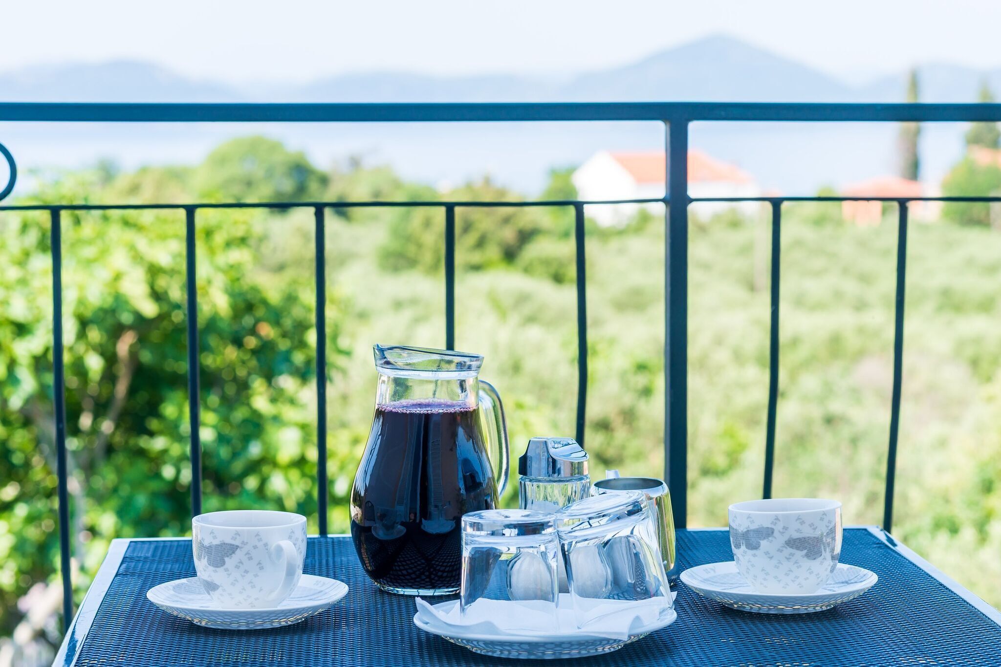 Ein Tisch mit einer Karaffe, Tassen und Zuckerbeschlägen auf einem Balkon mit Blick auf Grün und Berge.