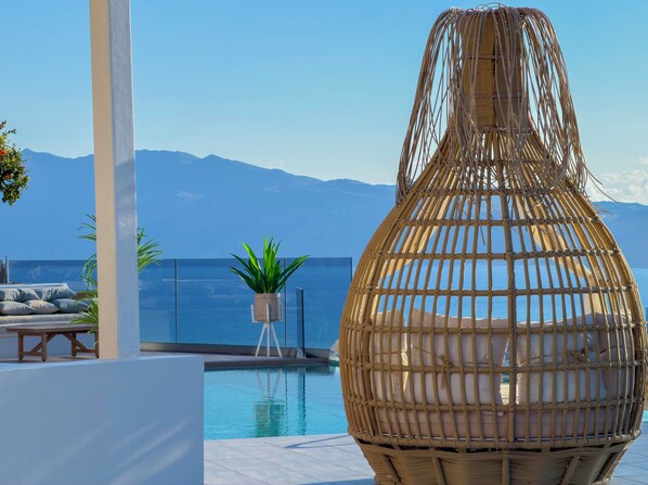 A pool area with a wicker chair and a potted plant, overlooking a body of water and mountains.