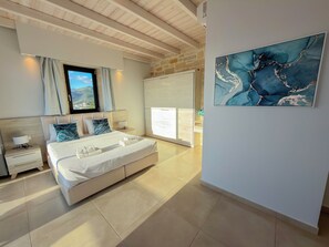A modern bedroom with a large bed, a nightstand, a window with curtains, a stone wall, and a framed abstract painting.