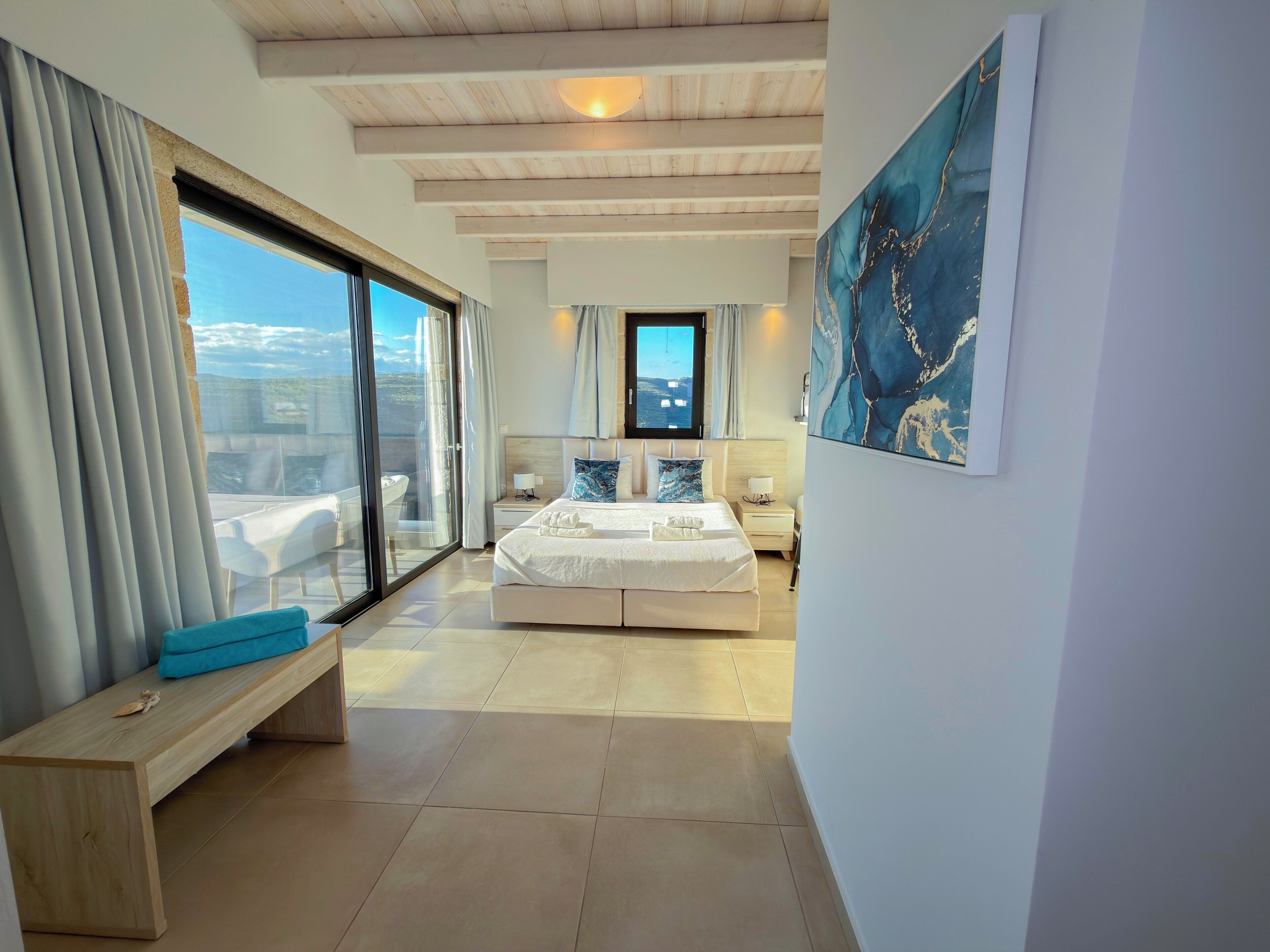 A modern bedroom with a large bed, a bench, a nightstand, and a view of the sea.
