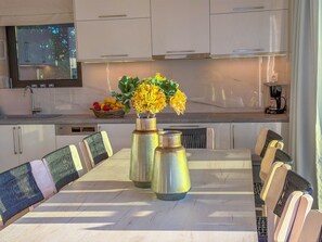 A modern kitchen with white cabinets, a dining table with chairs, a vase with flowers, and a fruit basket.