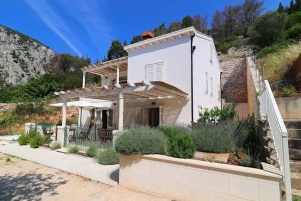 Una casa de dos pisos con un porche cubierto, una escalera de piedra y un jardín con diversas plantas.