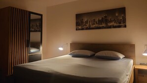 Un lit bien fait, avec deux oreillers, un tableau représentant un paysage urbain au-dessus, et une lampe de chevet de chaque côté.