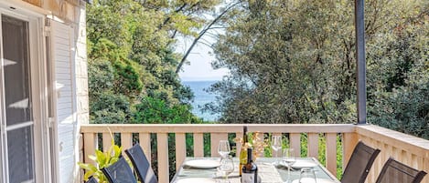 An outdoor dining area with a view of the sea, a table set with plates and glasses, and a bottle of wine.