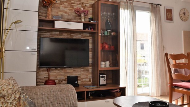Una sala de estar con televisor de pantalla plana, un mueble de madera con puertas de vidrio, una lámpara roja y una ventana con cortinas.