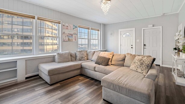 A modern living room with a sectional sofa, large windows, a chandelier, and a white door. - Glamorous Getaway - Steps to Beach! (Atlantic City)
