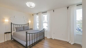 Een slaapkamer met een bed, nachtlampjes, een raam met gordijnen en uitzicht op gebouwen.