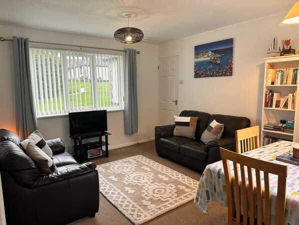 A living room with a black leather sofa, a wooden chair, a TV on a stand, a bookshelf, and a painting on the wall. - Gold Rated Modern Lodge With High Speed Wifi (Penzance)