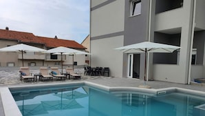 Una piscina con sillones y sombrillas, rodeada por un edificio con balcones y un techo de tejas.