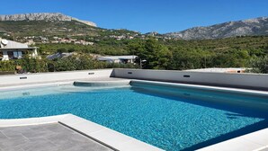 Una piscina con agua azul cristalina, rodeada por una solana con baldosas. Al fondo, se ven casas y una montaña.