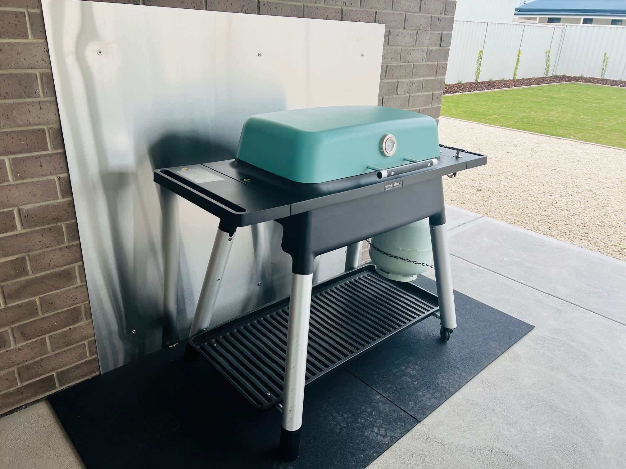 A portable gas grill with a black and teal color scheme, placed on a black mat outdoors.