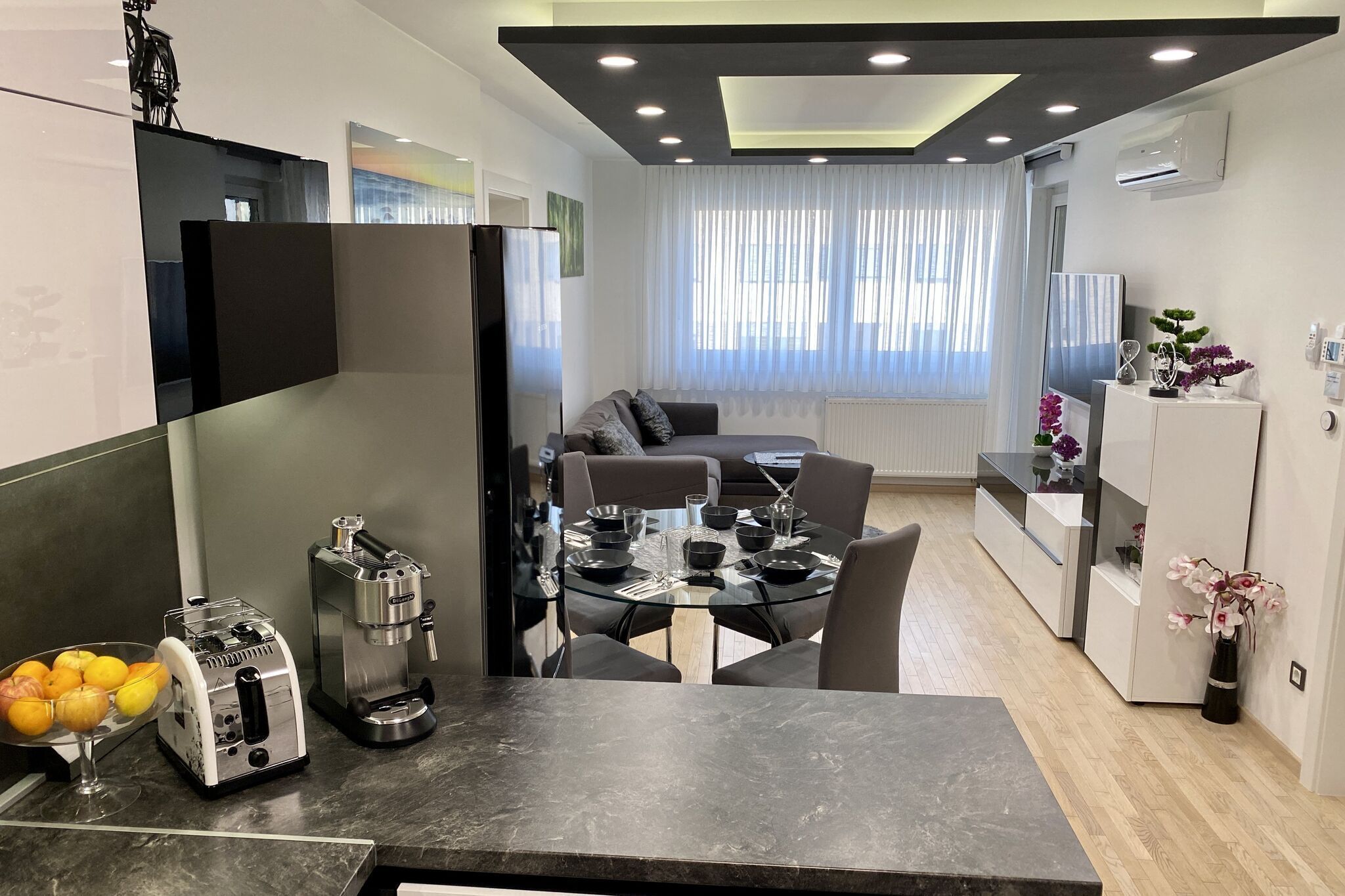 A modern kitchen with a dining area, featuring a coffee machine, fruit bowl, and a glass table.