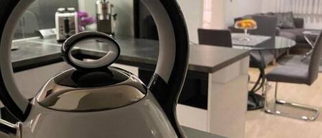 A modern kitchen with a kettle on a counter, a dining area with a table and chairs, and a sofa in the background.