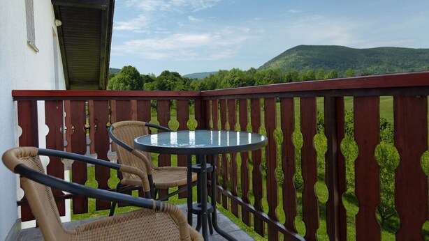 A balcony with wicker chairs and a glass table, overlooking a green landscape with hills. - Guesthouse Žafran- Room With Landscape View (Plitvicka Jezera)