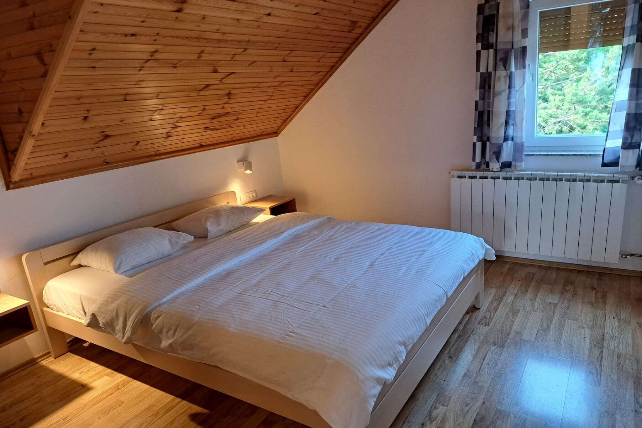A bedroom with a sloped ceiling, a bed with white bedding, a bedside table, a window with curtains, and a radiator.