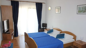 Uma cama de casal com roupa de cama azul, uma escrivaninha de madeira com televisão, uma pequena mesa de cabeceira e uma janela com cortinas.