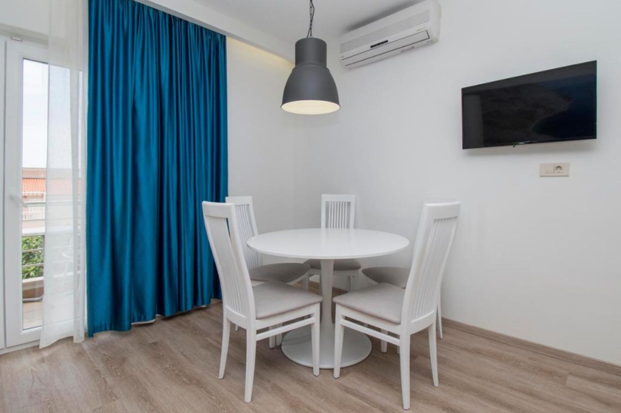 A room with a round white table and four white chairs, a black pendant light, a flat-screen TV on the wall, and a large window with blue curtains.