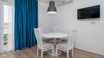 A room with a round white table and four white chairs, a black pendant light, a flat-screen TV on the wall, and a large window with blue curtains.