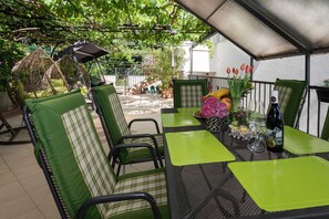 An outdoor patio with green cushioned chairs and a table set for dining, surrounded by plants and a pergola.