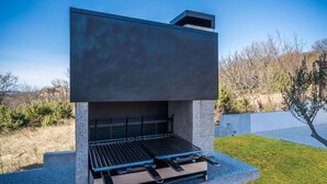 Een moderne buitenbarbecue met een vlakke plaat en een geïntegreerde plank, gelegen voor een heldere lucht en omgeven door een verzorgd gazon.