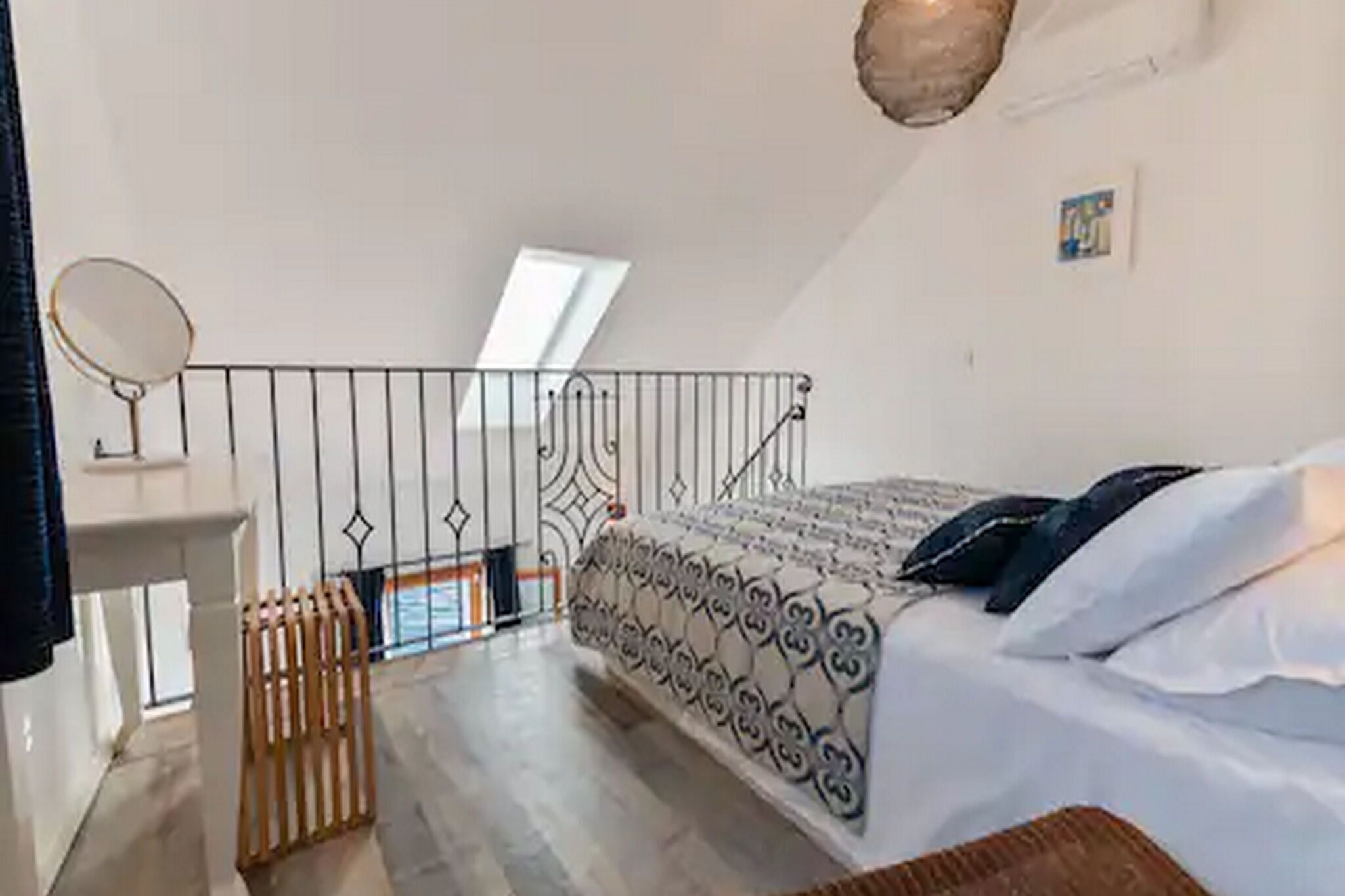 A bedroom with a bed, a small table with a mirror, a window, and a balcony railing.