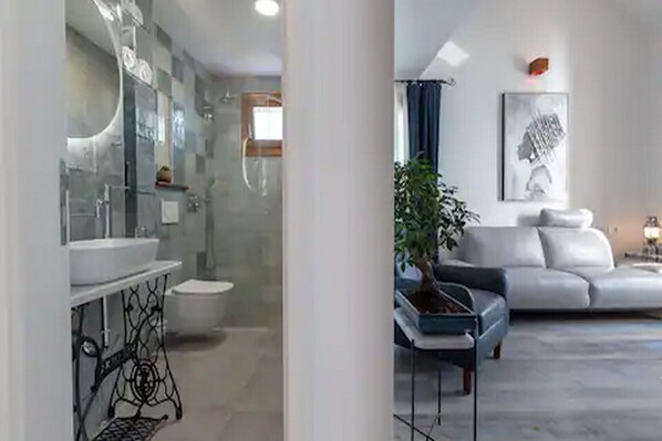 Een moderne badkamer met een douchecabine van glas, een witte wastafel en een toilet. In de woonruimte staat een witte bank en een klein tafeltje met een plant.
