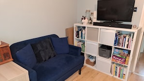 A living room with a blue sofa, a bookshelf filled with books, a television, and a small decorative pig figurine. - Garden-parking-3 Guests-wifi-20minsgatwick (Horsham)