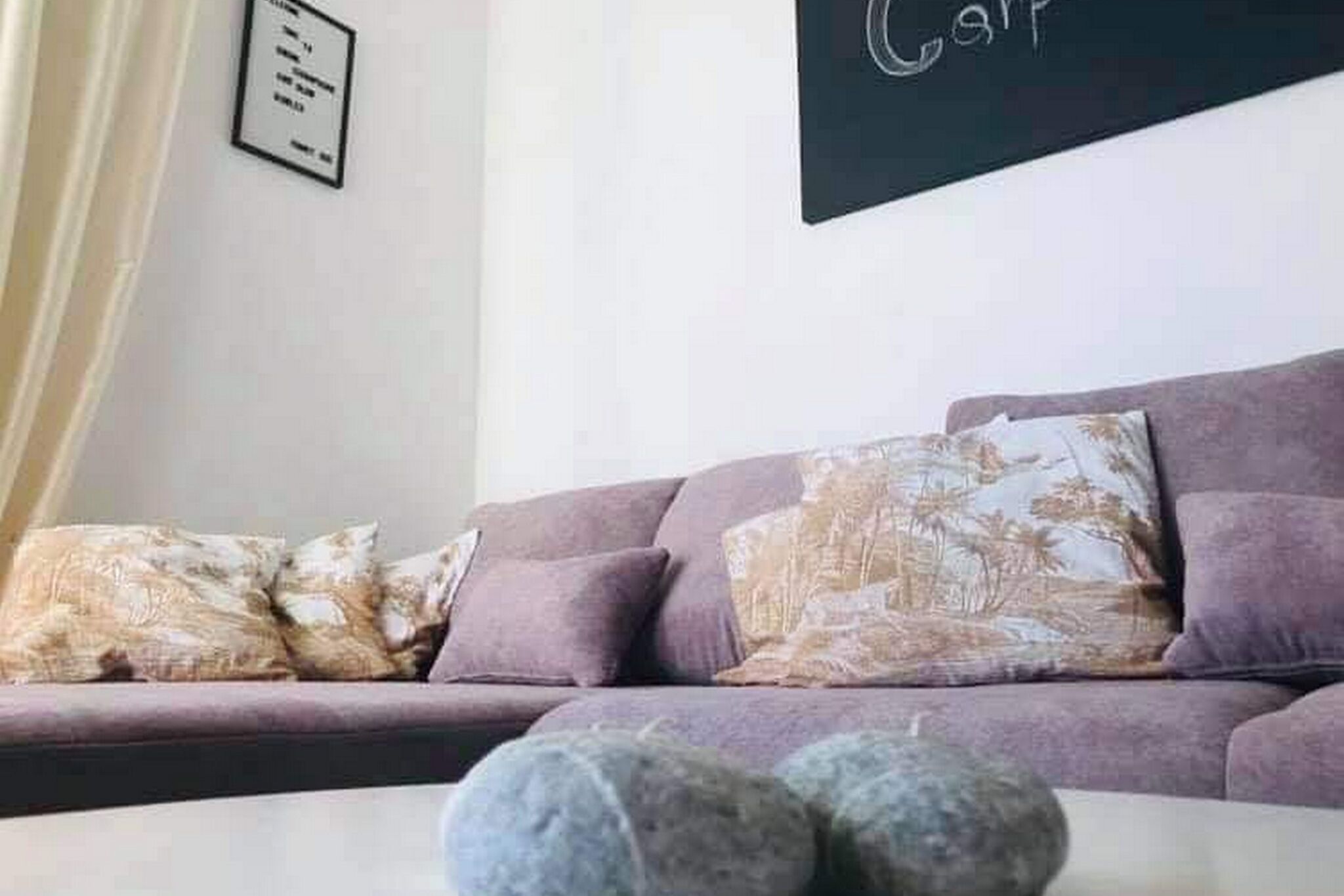 A purple sofa with patterned cushions, a framed sign on the wall, and a white table with two round objects.