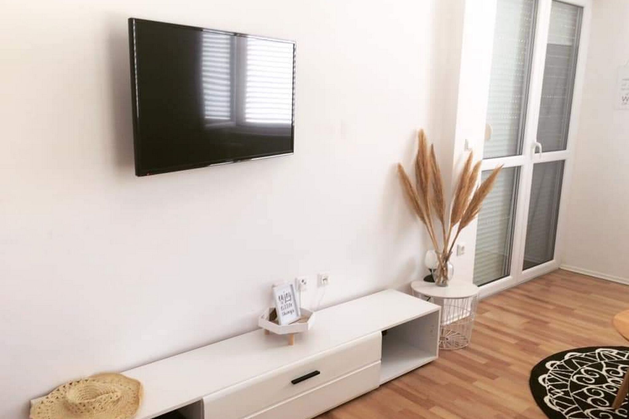 A modern living room with a flat-screen TV mounted on the wall, a white console table with a small basket, and a large window with blinds.